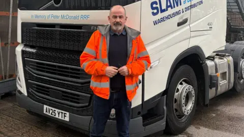 A man wearing an orange high-visibility jacket stands in front of a white Volvo lorry cab branded with "Slam Transport of Coventry - Warehousing & Distribution". The truck is parked on a wet concrete yard beside a large grey industrial warehouse. The man has a grey beard and is wearing dark jeans and brown boots, with his hands clasped together in front of him.