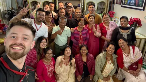 We Hate The Cold Harry Jackson pictured with a large group of people, in Nepal. They are in a room, all smiling and looking at the camera. Some women are sitting or kneeling and the rest are standing up. Many have their thumbs up. 