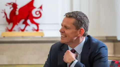 Rhun a Iorwerth sitting down, wearing a dark suit sits at a table, with a large red Welsh dragon emblem visible in the background of a light-coloured room.