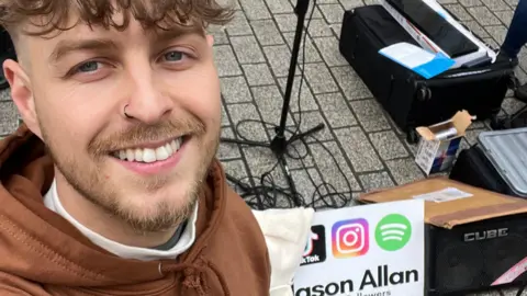 A man with brown hair and a small beard, with a black nose ring, wearing a brown hoodie smiles at the camera. He is standing next to music equipment.