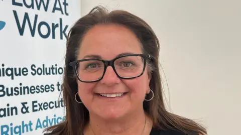 Heidi Gibaut is a woman with straight brown shoulder-length hair, medium-sized silver hoop ear-rings and large black-rimmed glasses. She smiles at the camera while stood in front of a white wall and a poster for her business.