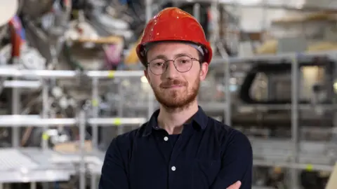 Proxima Fusion Wearing a red safety helmet and round glasses, Francesco Sciortino stands in front of fusion equipment.