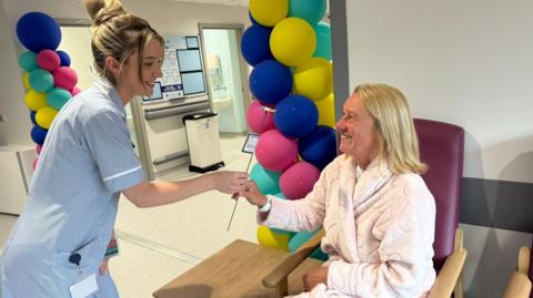 Jayne Hunt, sitting in a hospital chair, smiling and wearing a dressing gown, is treated by a nurse in front of balloons marking the opening.