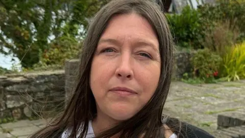 A woman with long dark brown hair is looking at the camera. She is wearing a white top with a black cardigan. She is standing on a paved area with trees and the sea blurred out behind her.