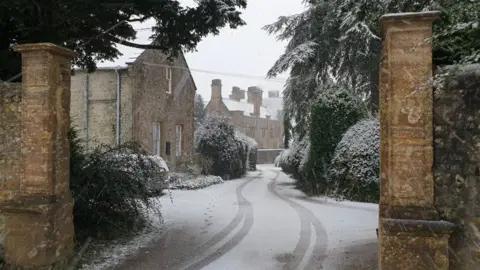 Stone gated entrance looking along a snow covered road and buildings.