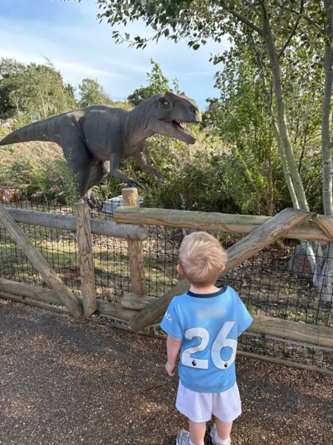 Charli Aitken Rudi, dressed in a light blue football top with the number 26 on the back and white shorts. He is looking at a large-scale model of a dinosaur in a park. 