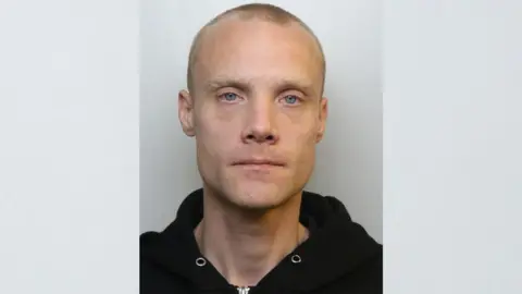 Custody photograph of Mitchell Lee Rogers. He has blonde hair that is shaven, blue eyes and a large visible scar on his left cheek. He is wearing a black hooded jumper with the hood down.