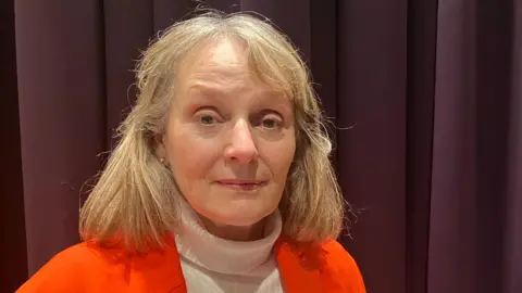 Amy Holmes/BBC A picture of Jane Whild who has light-coloured, shoulder‑length hair. She is standing indoors in front of dark, floor‑length curtains. She is wearing a bright orange jacket over a white, high‑neck top. The lighting highlights the clothing and hair against the darker background.