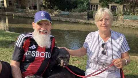 Dennis & Jackie King sit together on a grassy bank beside a quiet, tree‑lined river. Dennis is wearing a blue cap and a striped football shirt, Jackie has on a a white‑and‑blue striped top with sunglasses hanging from the neckline. Between them is a small black and grey dog on a red lead.