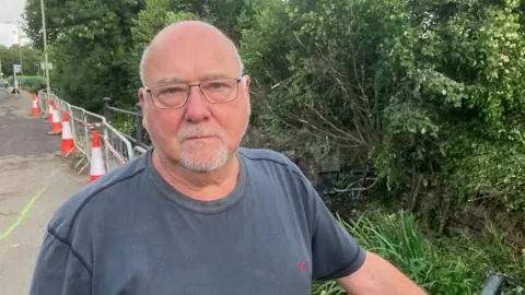 Dr John Fisher standing with his arm on a railing by a road crossing over a river - he is bald with glasses and is wearing a grey T-shirt.