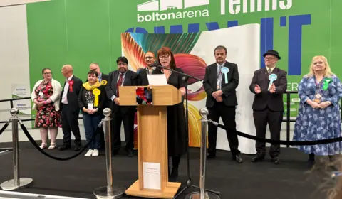 LDRS A woman with red hair stands speaking at a lecturn on a stage. Nine men and women stand behind her. 