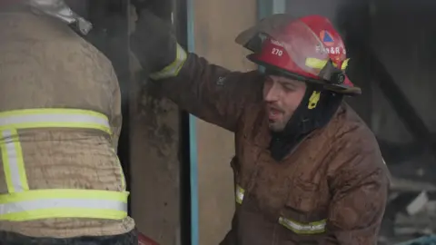 Civil Defence worker fights a fire. He is wearing a red helmet and looks breathless. 