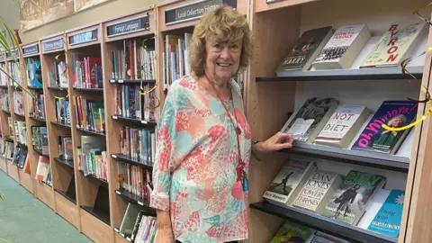 A woman in an aqua, pink and orange seashell print top with brown curly shoulder length hair, standing in a library