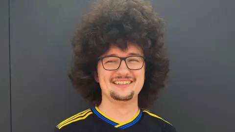 Frank Ilett/Instagram A head-and-shoulders image of Frank Ilett. He has voluminous curly dark hair and he is standing smiling against a grey backdrop.