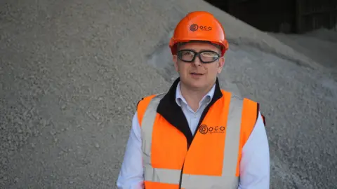 Jamie Niblock/BBC Graham Cooper in front of a pile of limestone aggregate. He wears an orange hard hat with O.C.O on it and an orange hi-vis vest over a blue and white pinstriped shirt. He has black rimmed goggles on.