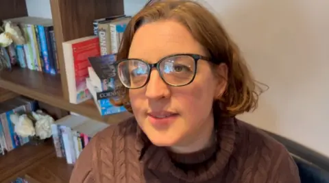 Martin Heath/BBC Annie Dupree with medium-length brown hair wearing brown-framed glasses and a brown sweater. She is sitting in a cafe with book shelves to her right.