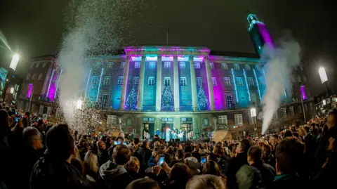 Norwich City Council Norwich City Hall, with its numerous large columns, lit up in blue and purple. Christmas trees adorn the balcony and hundreds of people are stood outside, many of them holding up their mobile phones.