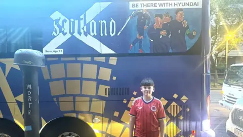 A boy wearing a red football top stands beside the Scotland World Cup team bus, which features his design