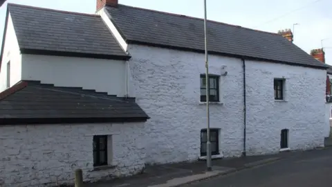 John Lord/Geograph The outside of a white-wash farm house with several windows in it.