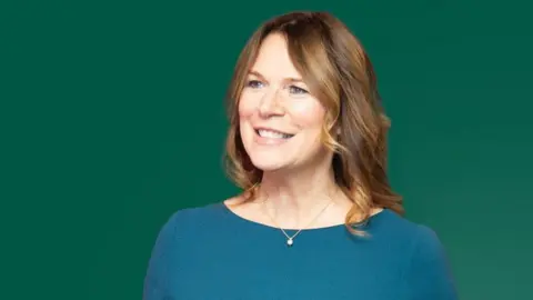 OurCoop A woman with shoulder-length brown hair, wearing a dark green top and a necklace, is seen smiling whilst looking off to her right. She is standing against a green background.