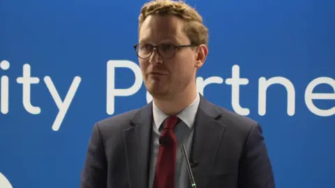 A man with glasses and brown hair is addressing an audience. He is wearing a shirt, dark jacket and red tie and standing in front of a mainly blue background. 