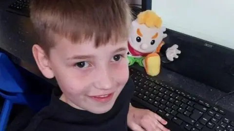 Family photo Leighton sitting at a computer is smiling at the camera with his puppet