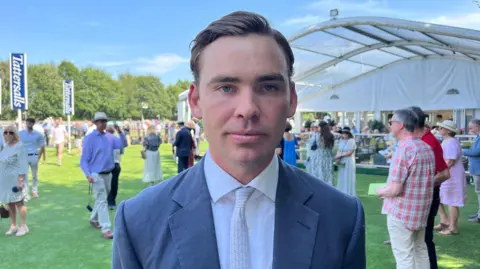 Tom Williams/BBC Thady Gosden standing outdoors at Newmarket Racecourse. He is looking directly at the camera and is wearing a grey blazer, white shirt and tie. There are groups of people walking around the course behind him.