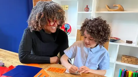 Christina Lipede at home with her son, aged nine. The pair are sitting at a table and her son is writing.