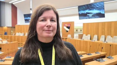 Paul Moseley/BBC Emma Corlett is looking at the camera. She's wearing a dark woolly top with a yellow lanyard. Behind her is a council chamber, decorated with wooden beech furnishings.