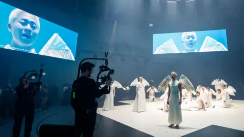 Tristram Kenton Press photograph of Angel's Bone at the Aviva Studios in Manchester. The image shows the cast on stage wearing angel constumes, a mobile camera and two giant screens in the background.