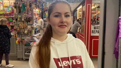 A woman with dark hair in a pony tale over one shoulder of a white and red hooded top stands in a market with a variety of stalls visible behind her
