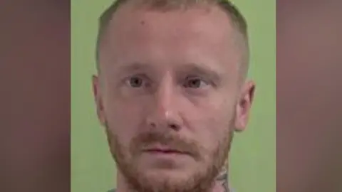A police mugshot of Fayle, who has short light brown hair and a close-cropped beard and a tattoo on his neck. He is standing against a green background.