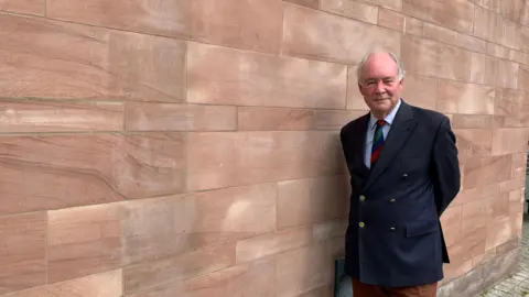The image shows a late middle-aged man with a balding head and grey hair at the sides, wearing a navy blue blazer over a light blue shirt with a green, blue and red striped tie, standing next to a wall of light red sandstone.