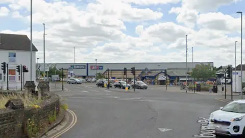 A Google Maps image of the junction with traffic lights, cars and people walking.