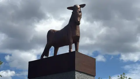 A bronze-coloured fibreglass statue of a pony stands fixed to a wooden board, which is on top of a concrete plinth.