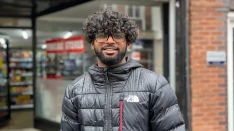 A man with black curly hair, glasses and black beard. He is wearing a black puffa jacket and is standing in front of a shop 