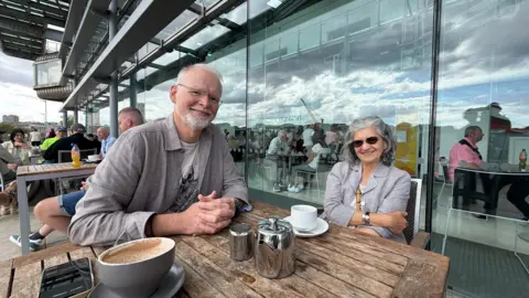 Jim Scott/BBC Stuart and Judith Crompton smiling into the camera while sitting at an outdoors coffee table in front of the glass building. Stuart is on the left and has white short hair and a white beard. He is wearing glasses and a grey shirt over a white graphic t-shirt and has his hands clasped in front of him on the wooden table. Judith is on the right and has grey, shoulder length hair and has clipped the front parts clipped back. She is wearing dark sunglasses and a grey blazer with the strap of a brown cross body bag visible. One of her arms is crossed over her stomach. There is a large coffee cup on the table as well as a silver tea pot, milk jug and white tea cup.