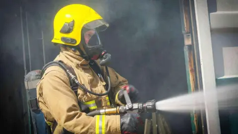 A firefighter in a yellow helmet and beige uniform shoots water from a hose to the right of the photo