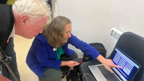 Hilary Jeune and Peter McLinton crouch down to look at a laptop screen which is on a black chair. Jeune is pointing at the screen with her left hand.