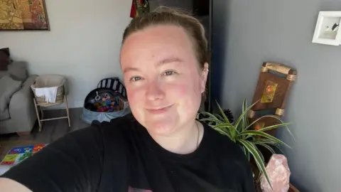 Anik Hubert Anik has her dark brown hair tied back in a ponytail. She is wearing a black t-shirt and smiling directly at the camera from her living room.