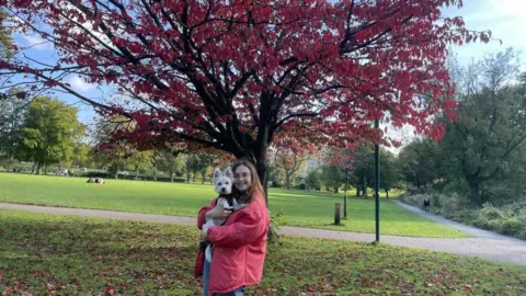Chloe Price Chloe Price, who is wearing a red jacket, is holding a dog. There is a tree in a red autumn colour behind her.
