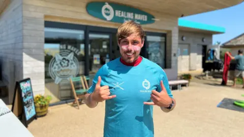 BBC A man in a blue T-shirt with sun block on his cheeks smiles at the camera as he stands in front of the Healing Waves office - a modern, wooden structure with large windows near a beach.