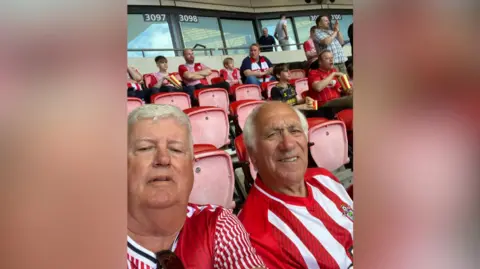 Ian Mcauley Two men sitting at a football stadium with football shirts on. They are smiling for a selfie. Other fans can be seen behind them. 