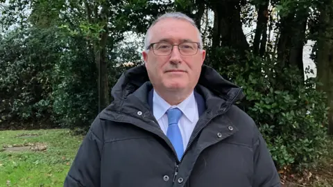 Shows Paddy MacDermott in a white shirt, blue tie, jacket and hooded coat wearing glasses and standing in front of trees