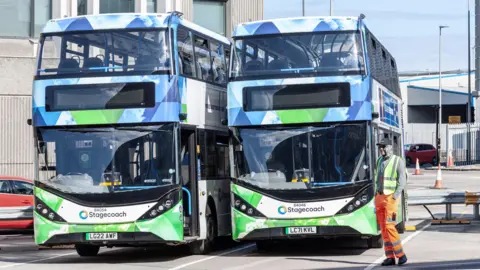 Two Stagecoach buses next to each other in a depot, while a bus worker stands nearby