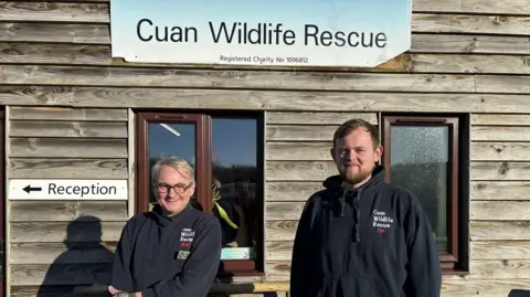 Two men are standing outside the Cuan Wildlife Rescue building. It is a wooden building with a white sign that reads "reception". The man on the left has grey hair and glasses, and the man on the right has brown hair and brown facial hair. They are both wearing Cuan Wildlife Rescue branded navy hoodies.