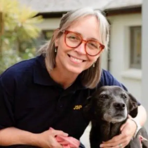 JSPCA Pam Aubert smiling at the camera while cuddling a black dog. She is wearing glasses, earrings, a navy blue polo shirt and has short blonde hair.