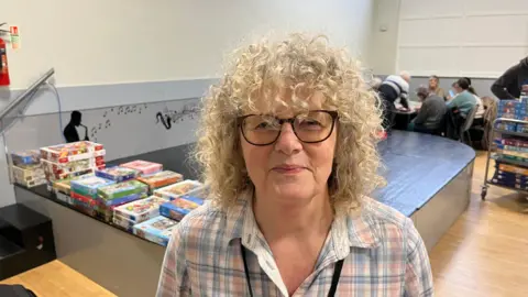 John Devine/BBC Ruth Freeman has very curly blonde hair and dark framed glasses on, she has a light pink and blue tartan patterned shirt on, behind her is a stage with some jigsaw puzzle boxes on.