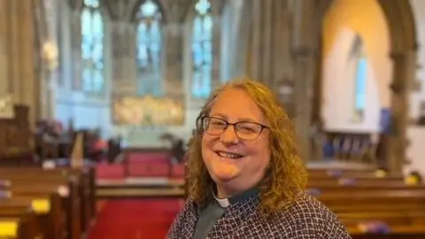 Reverend Heather Atkinson A smiling Reverend Heather Atkinson stands in the middle of her church. The altar and ornate stained glass windows stand behind her. She has curly blonde hair and glasses and is wearing a grey top with white dog collar and a purple-and-white patterned jacket.