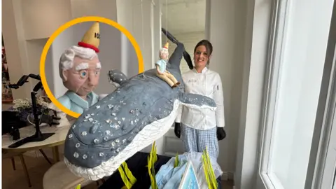 Female baker in front of a giant cake in the shape of a whale.